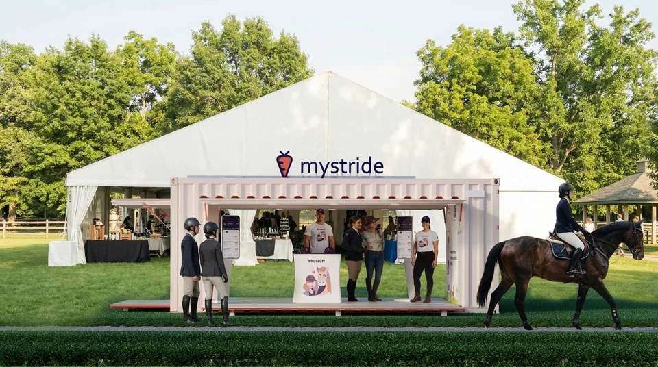 Front view 3D render of the MyStride container booth with its side door raised as a canopy, in front of a marquee at a horse show